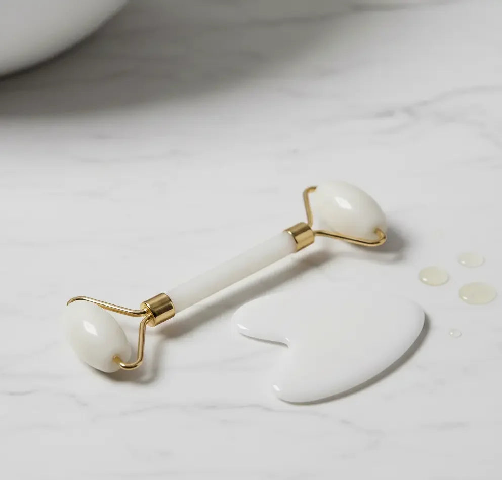White jade facial roller and gua sha stone on marble surface with skincare serum drops
